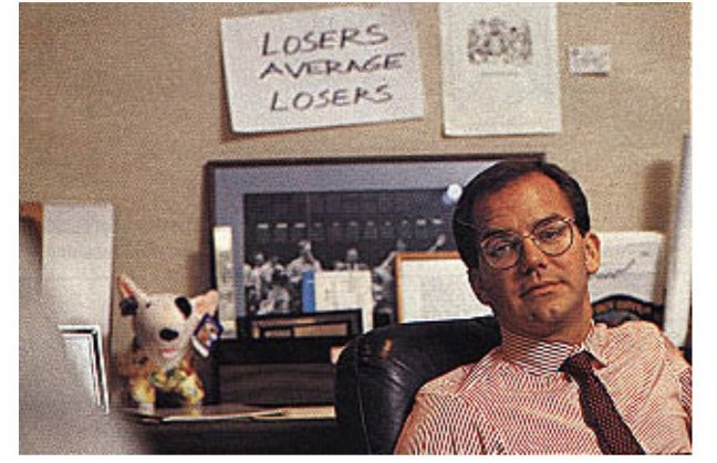 Paul Tudor Jones in glasses and a red-striped shirt sits in an office chair, with a focused expression. Behind him, a sign reads "LOSERS AVERAGE LOSERS." Nearby are framed pictures and a dog figurine, suggesting a professional yet quirky environment.