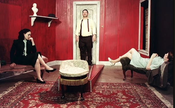 Three souls. One room. No escape. Three people in a red room: a man stands by a door, a woman in black sits thoughtfully, and another woman reclines on a couch. The setting is theatrical.