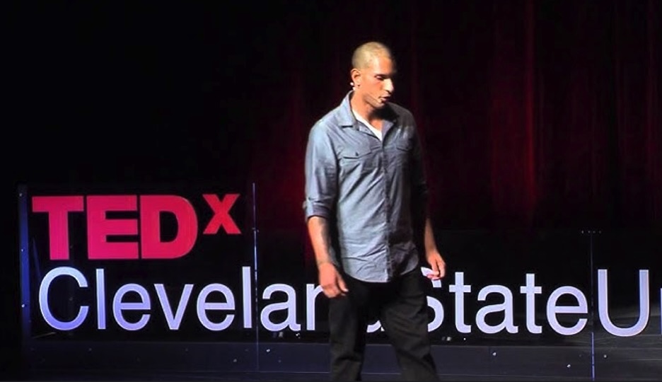 A speaker on stage at TEDx Cleveland State University, wearing a blue button-up shirt. The background is a dark red curtain, and a TEDx sign is visible.
