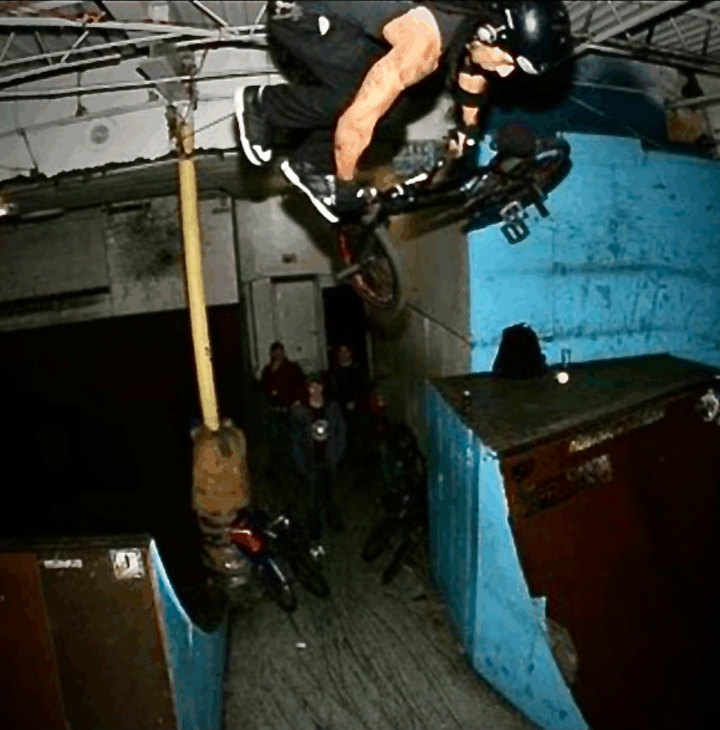 A BMX rider in a black helmet performs an aerial trick in an indoor skate park with blue ramps. Onlookers watch from below, adding a sense of excitement.