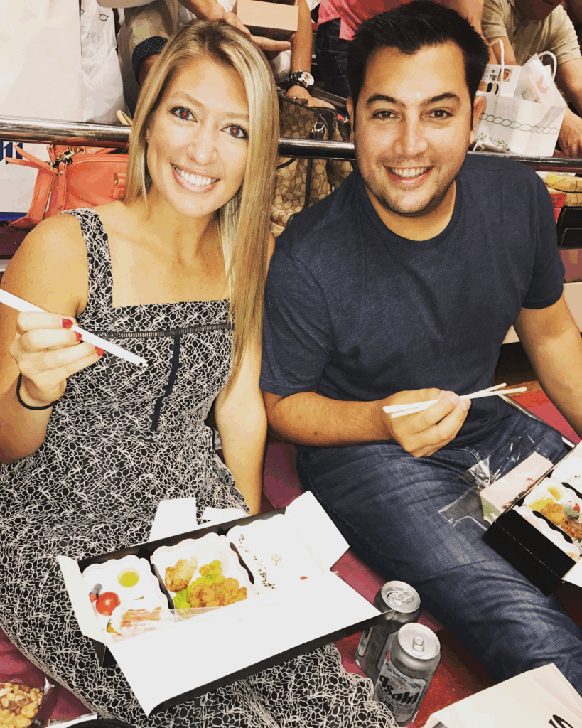 A smiling woman and JC sit closely at the premier Sumo stadium, holding chopsticks and enjoying boxed meals.
