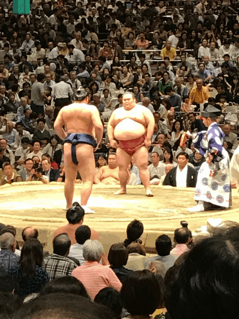 Two sumo wrestlers face off in a crowded arena. One wears a blue mawashi, the other red. A referee in traditional attire stands nearby, observing. Spectators fill the background.