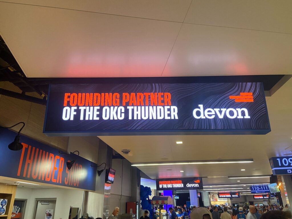 "Founding Partner of the OKC Thunder" with "devon" logo. Located inside an arena with people shopping below at the Thunder Shop.